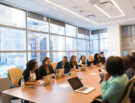 Business meeting table with roughly fourteen people - who are mostly black - sitting around it with their respective laptops or tablets.