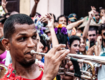 [Homem preto de cabelo raspado, camiseta vermelha em destaque no centro da imagem, tocando um trompete. Ao seu redor, várias pessoas com fantasias e os rostos pintados para o Carnaval.]