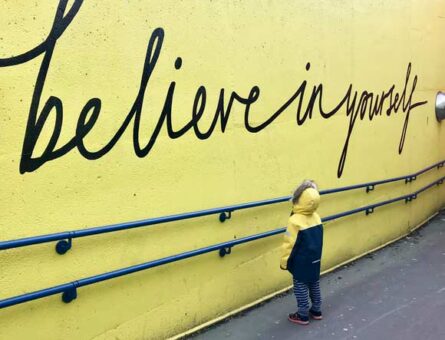 Parede amarela com o texto “believe in yourself”, que significa acredite em si mesmo em português. À frente da parede, há uma criança olhando para o escrito.