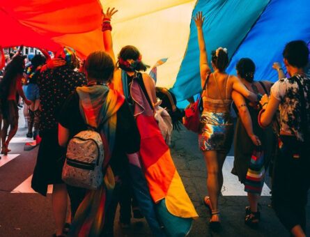 Dozens of LGBTQIA+ people under a rainbow flag