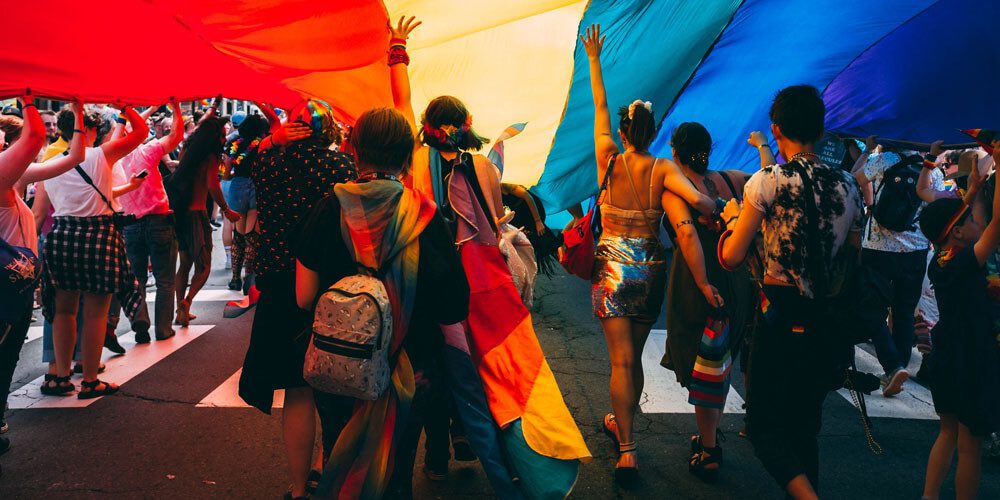 Dozens of LGBTQIA+ people under a rainbow flag