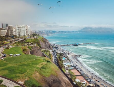 Aerial picture of the seaside of Lima, Peru. The beach has clear blue water and the coast has hills with white buildings on top of them.