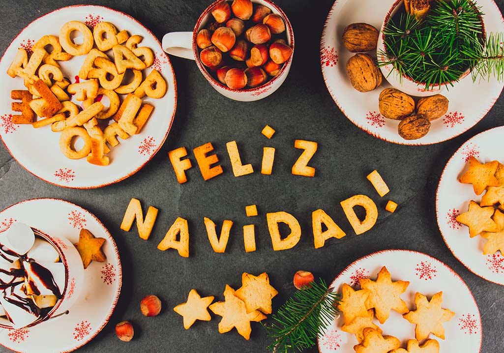 Words Merry Christmas in Spanish with baked cookies, Christmas decoration and nuts on black slate background.