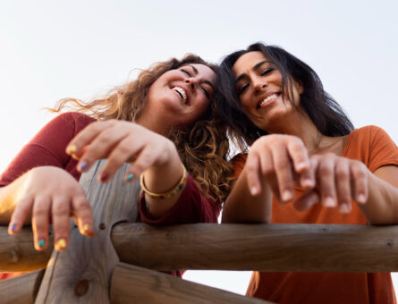 Dos chicas sonriendo para una foto, de pie una al lado de la otra. Ambas chicas visten ropa casual y tienen expresiones alegres y brillantes. El fondo es al aire libre, con luz natural, lo que crea una atmósfera cálida y amigable.