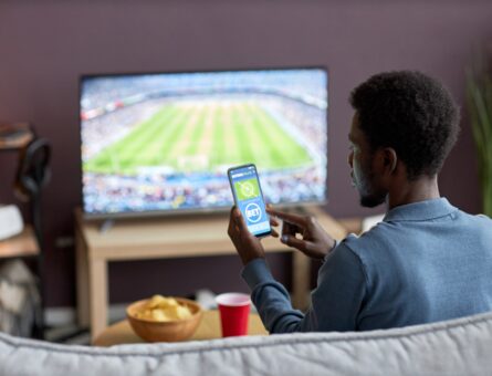 Back view of man holding smartphone with sports bets app on screen while watching football match at home, copy space